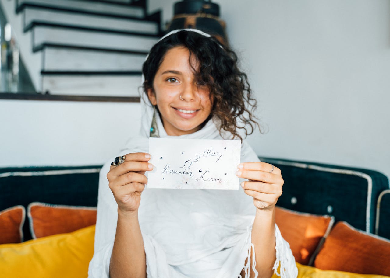 A joyful woman in traditional attire holds a Ramadan greeting card indoors.